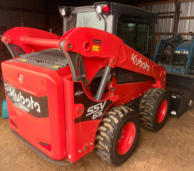 Roter Kubota SSV65 Radlader vor einer Scheune geparkt, mit anderen Fahrzeugen im Hintergrund.
