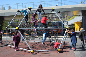Eine Gruppe von Kindern spielt auf einer Spielplatzstruktur vor einem Gebäude, umgeben von Bäumen unter einem klaren blauen Himmel, wobei einige Kinder stehen, auf Bänken sitzen oder um die Struktur herumlaufen.