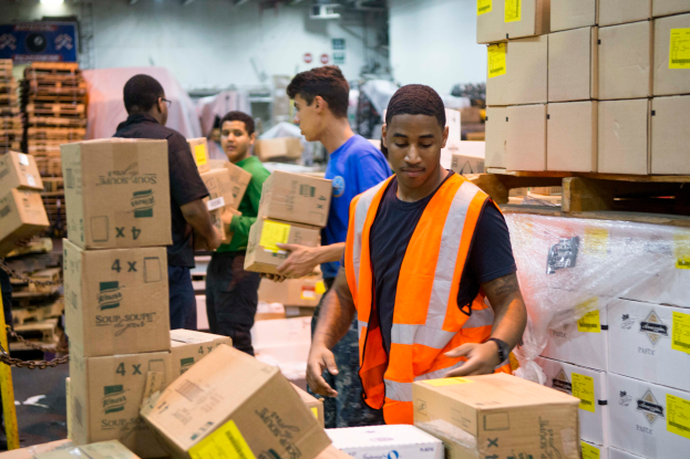 Eine Gruppe von Männern in orangefarbenen Westen und Sicherheitswesten lädt Kisten in ein Lagerhaus, mit mehreren Kartonagen auf der rechten Seite und einer Kette auf der linken Seite, und einer Wand mit Lampen im Hintergrund.