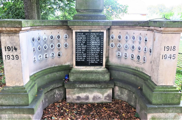 Ein Holocaust-Gedenkmonument in einem jüdischen Friedhof in Berlin, mit einer Wand voller Text und Zahlen, umgeben von Bäumen, einem Zaun und verstreuten trockenen Blättern.