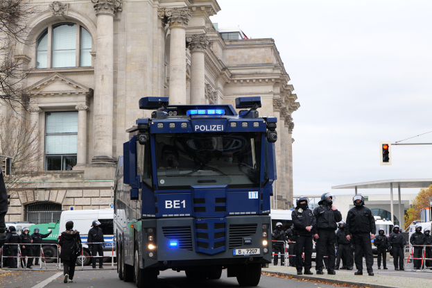 Eine Gruppe von Polizisten in Helmen steht vor einem großen Gebäude mit Fenstern, Säulen und Bögen, umgeben von einem Metallzaun, mit Fahrzeugen auf der Straße, einer Ampel, Bäumen und einem klaren blauen Himmel im Hintergrund.