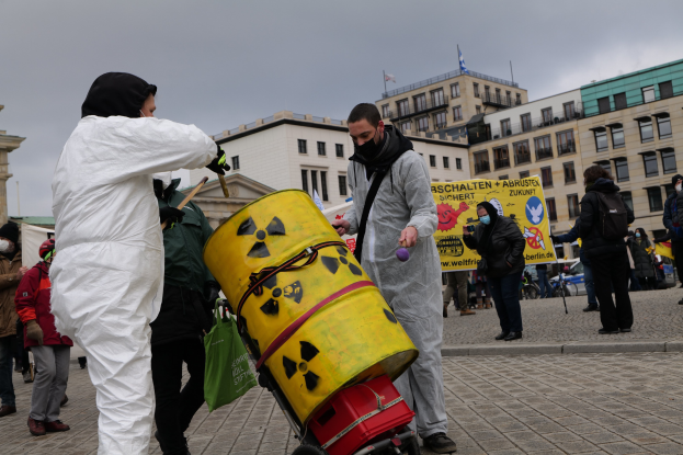 Eine Gruppe von Menschen in weißen Anzügen und Masken steht um einen gelben Eimer mit einem Karren im Vordergrund, hält Schilder in der Nähe von Gebäuden mit Fenstern und Flaggen in Berlin.