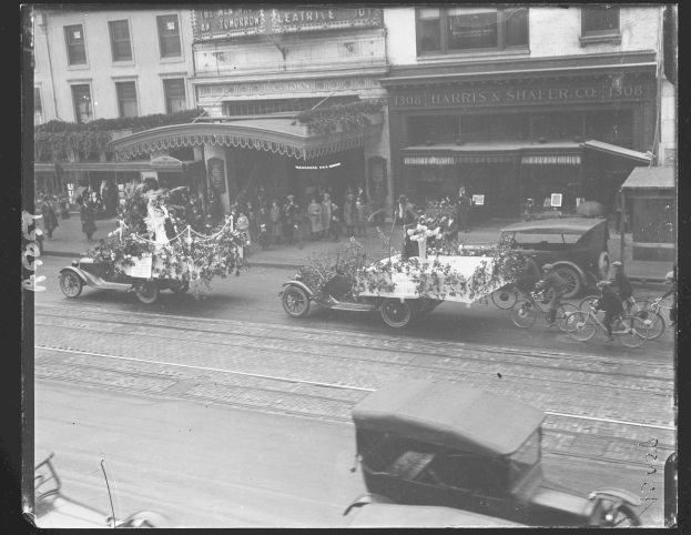 Ein Schwarz-Weiß-Foto eines Umzugs auf einer Stadtstraße mit Fahrzeugen, Passanten und Gebäuden im Hintergrund.