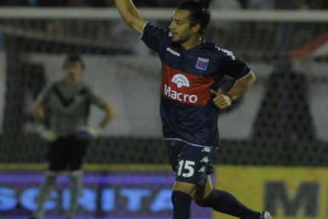 Ein Fußballspieler feiert nach einem Tor auf dem Boden, mit jubelnden Zuschauern und einer Person im Hintergrund und einer Tafel mit Text dahinter, alles vor einem leicht unscharfen Hintergrund.