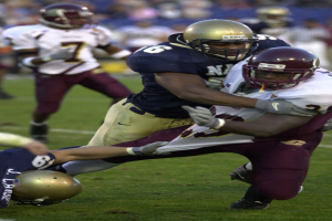 Zwei Fußballspieler in Helmen und Uniformen stoßen auf dem Feld zusammen, während andere zuschauen.
