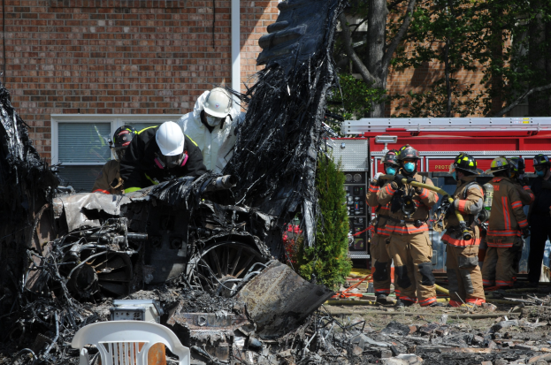 Feuerwehrleute in Schutzausrüstung löschen ein Gebäude Feuer, mit verkohlten Trümmern einschließlich eines Fahrzeugs, Stuhl und Pflanzen sichtbar auf dem Boden.