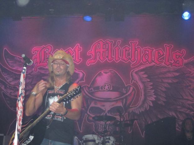 Eine musikalische Konzertsituation mit Scheinwerfern oben, einem Banner im Hintergrund, Drogen in der Mitte unten, einer Person in der rechten unteren Ecke und einem Gitarristen mit Mikrofon links.