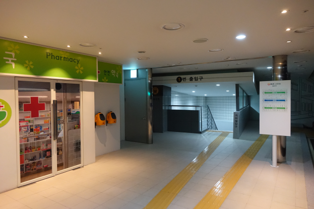 Der Eingang einer Apotheke mit einem 'Apotheke'-Schild auf der linken Seite, einer Tafel mit Text auf der rechten Seite und beleuchteten Lichtern auf dem Dach.