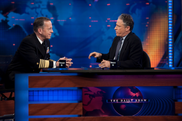 Zwei Manner in schwarzen Blazern, Hemden und Krawatten sitzen an einem Tisch mit Mikrofonen zwischen ihnen und fuhren ein Gesprach vor einem Fernseher und einem blauen Hintergrund.