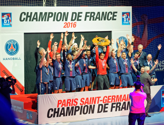 Männergruppe auf einer Bühne mit einem Pokal, einer Person filmt und einem "Champion de France 2016"-Schild im Hintergrund.