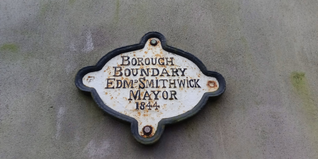 Metallplatte an der Außenseite eines Gebäudes mit schwarzem Hintergrund und weißer fettgedruckter Schrift, die "Gemeindegrenze Edm Smithwick Bürgermeister" lautet.