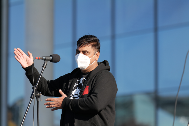 Ein Mann in einer schwarzen Kapuzenjacke und Gesichtsmaske steht vor einem Mikrofon, mit einem unscharfen Hintergrund.