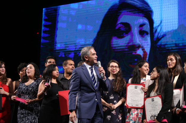 Ein Mann im Anzug und Krawatte steht auf einer Bühne mit einem Mikrofon, umgeben von Menschen mit Urkunden, mit einem Bild einer Frau im Hintergrund.