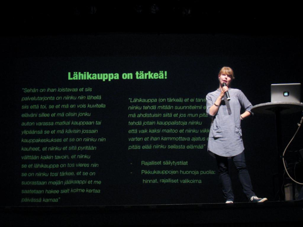 Eine Frau steht auf einer B├╝hne und spricht, mit einem Apple-Laptop auf einem Tisch neben ihr und einem Projektor, der hinter ihr Inhalt anzeigt.