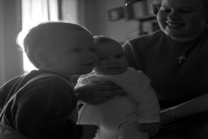 Eine Frau lächelt warm, während sie ein Baby hält, mit zwei anderen Kindern, die neben ihr stehen, in einem gemütlichen Raum mit Rahmen und einem Fenster im Hintergrund.