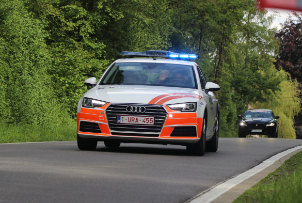 Ein Audi A4 Polizeiwagen fährt auf einer Straße neben einem Wald, mit zwei Personen darin, Gras auf beiden Seiten, Bäumen und einem klaren blauen Himmel im Hintergrund.