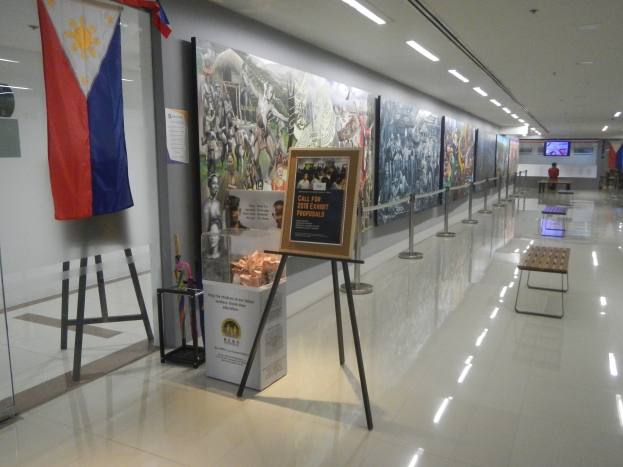 Langer Flur mit einer philippinischen Flagge an der Wand, Displaytafeln mit Text und Bildern, Ribbonständer, Bänke, Deckenleuchten und ein Bildschirm im Hintergrund, der Museumsinformationen zeigt.