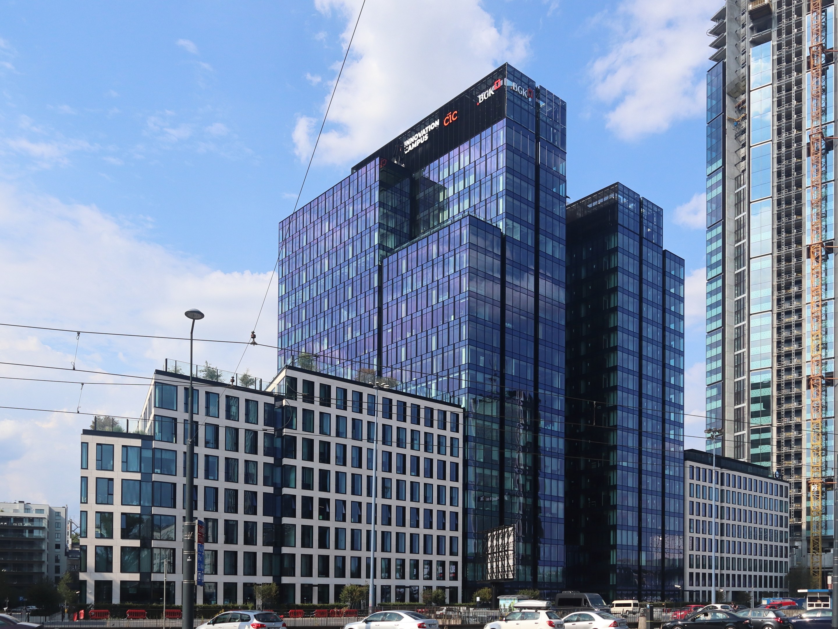 Großes modernes Gebäude mit vielen Fenstern, identifiziert als Deutsche Bank Hauptquartier in Frankfurt, Deutschland, umgeben von städtischer Infrastruktur einschließlich Straßenlaternen, Lichtern, Fahrzeugen, Grünflächen und einem bewölkten Himmel.