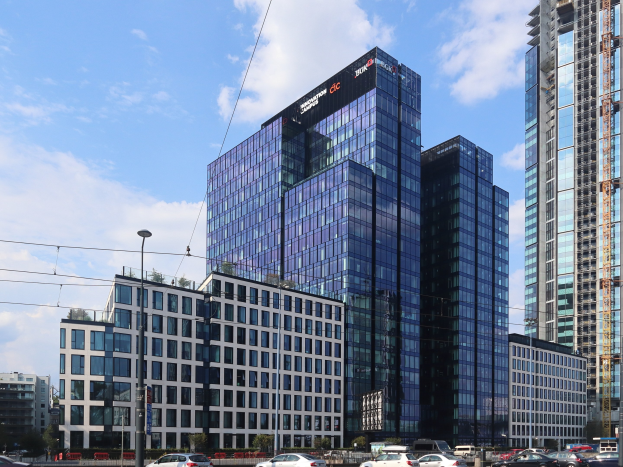 Großes modernes Gebäude mit vielen Fenstern, identifiziert als Deutsche Bank Hauptquartier in Frankfurt, Deutschland, umgeben von städtischer Infrastruktur einschließlich Straßenlaternen, Lichtern, Fahrzeugen, Grünflächen und einem bewölkten Himmel.