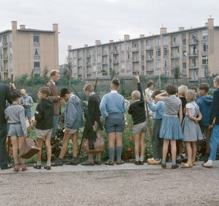 Kinder vor einem Gebäude mit Bäumen und Blumen, einige halten Schulranzen unter einem klaren blauen Himmel.