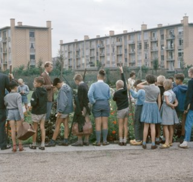 Kinder vor einem Gebäude mit Bäumen und Blumen, einige halten Schulranzen unter einem klaren blauen Himmel.