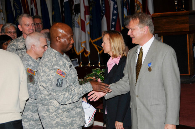 Eine Gruppe von Menschen, die mit zwei Personen stehen, die Hände schütteln, Flaggen im Hintergrund, ein Podium mit einem Mikrofon und eine Topfpflanze in der Nähe.