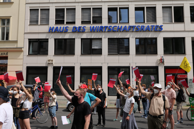Eine Gruppe von Menschen, die vor einem Gebäude protestiert und Schilder und Fahnen schwingt, mit Fahrzeugen auf der Straße und einem Schild auf der rechten Seite.