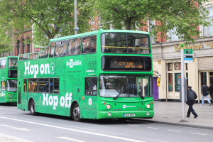 Zwei grüne Doppeldeckerbusse fahren auf einer Straße mit hohen Gebäuden, Menschen auf dem Bürgersteig, einem Schild, Bäumen und einem klaren blauen Himmel im Hintergrund.