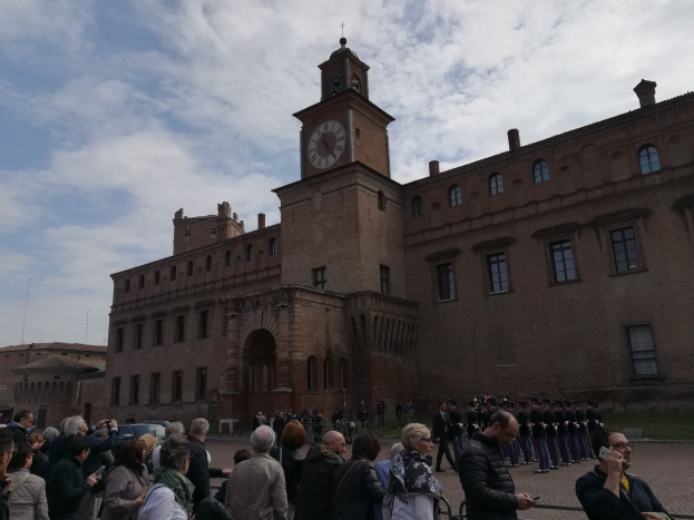 Eine Gruppe von Menschen vor einem großen braunen Gebäude mit einem Uhrenturm, einige halten Mobiltelefone, unter einem blauen und weißen Himmel.