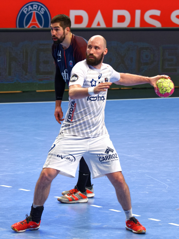 Zwei Männer spielen Handball auf einem Platz, einer hält den Ball, im Hintergrund ein Schild mit der Aufschrift "Paris Saint-Germain vs Paris Saint-Germain."