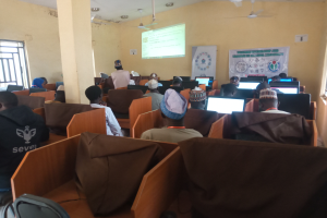 Eine Gruppe von Menschen, die auf Stühlen sitzen und Computern in einem Raum mit einem Bildschirm, einer Fahne, Deckenventilatoren und Fenstern auf beiden Seiten gegenüber sitzen und an einem Seminar zur Technologieentwicklung in Nigeria teilnehmen.