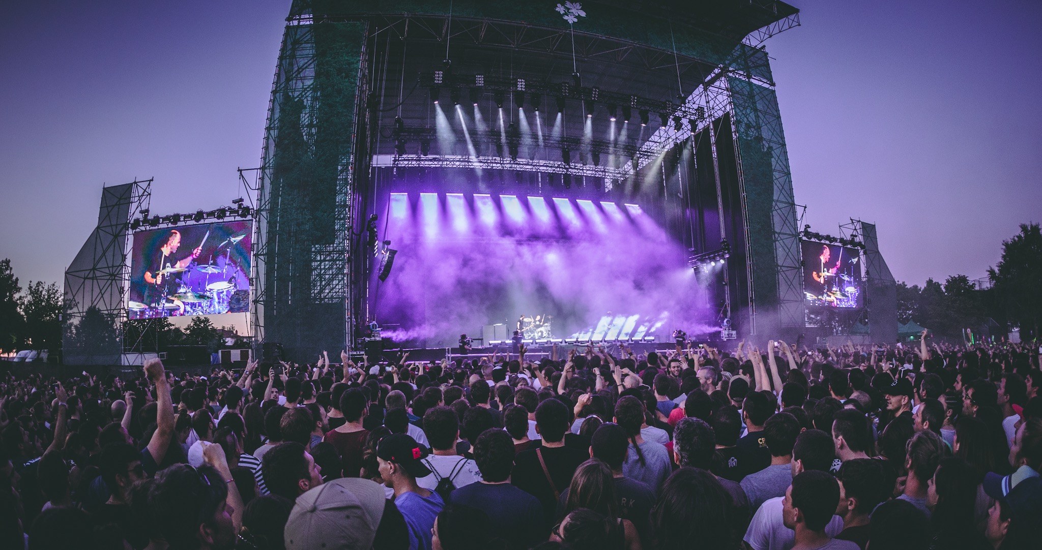 Eine große Menschenmenge auf einem Musikfestival schaut auf der beleuchteten Bühne mit Instrumenten und Bildschirmen, die vor einem Gebäude, Bäumen und einem klaren blauen Himmel aufgebaut ist.