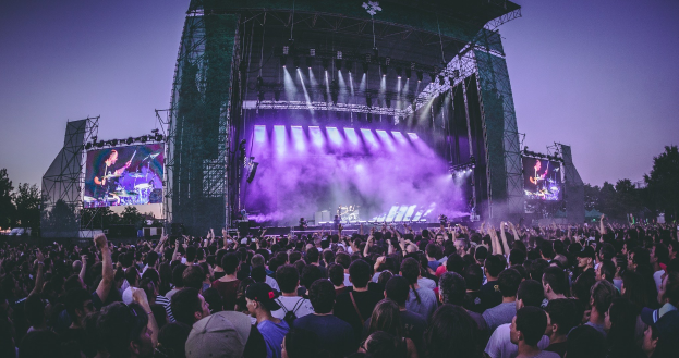 Eine große Menschenmenge auf einem Musikfestival schaut auf der beleuchteten Bühne mit Instrumenten und Bildschirmen, die vor einem Gebäude, Bäumen und einem klaren blauen Himmel aufgebaut ist.