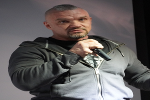 Ein Mann in einer schwarzen Kapuzenjacke und einem schwarzen Anzug mit weißem Hemd steht selbstbewusst vor einer Wand und hält ein Mikrofon, wobei er wie ein Komiker wirkt.