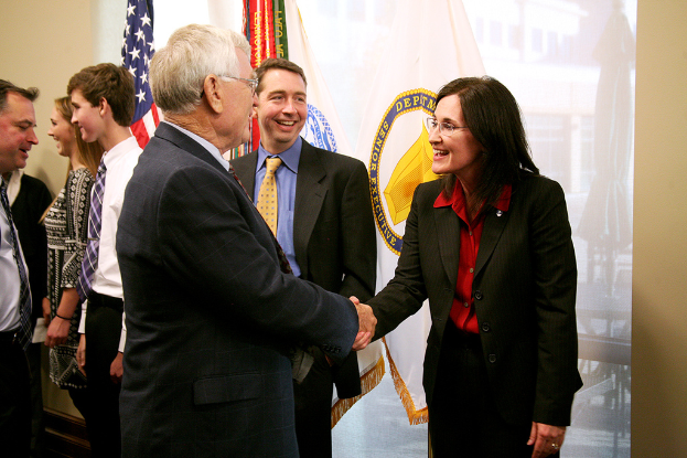 Ein Mann und eine Frau in Blazern und Hemden geben sich die Hand und lächeln sich an, während ein Mann in Blazer, Krawatte und Hemd neben ihnen steht; im Hintergrund unterhalten sich Menschen und lächeln mit sichtbaren Fahnen an der Wand.