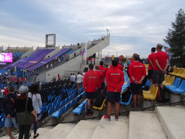 Eine Gruppe von Menschen, einige sitzend und andere auf einer Treppe stehend, versammelt vor einem Stadion mit einem Baum auf der rechten Seite, einem Bildschirm auf der linken Seite, Laternenmasten und einem klaren blauen Himmel im Hintergrund.