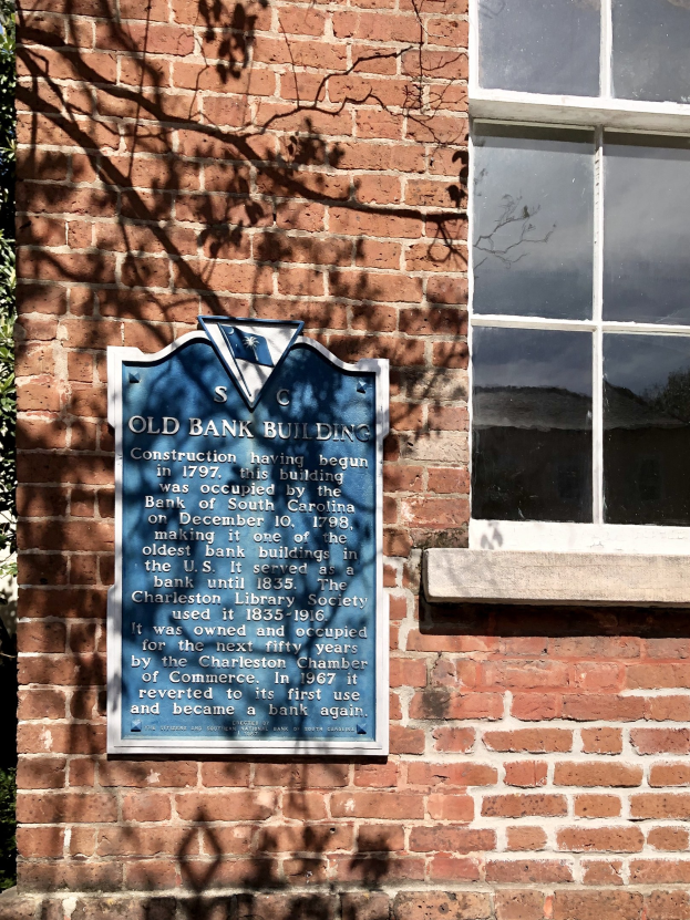 Altes Bankgebäude mit blauer Plakette an der Seite, einem Fenster rechts und einem Baum links.