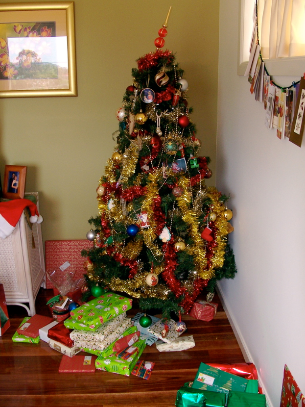 Ein geschmückter Weihnachtsbaum mit Geschenken am Boden, ein Photo frame an der linken Wand und ein Vorhang am rechten Fenster.