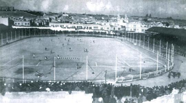 Ein Schwarz-Weiß-Foto eines Fußballfeldes mit einer Gruppe von Menschen, die in der Mitte Fußball spielen, umgeben von Pfählen und einem Zaun, mit Gebäuden und einem bewölkten Himmel im Hintergrund und einer Abfolge von Jahren von 1908 bis 1993 unten.