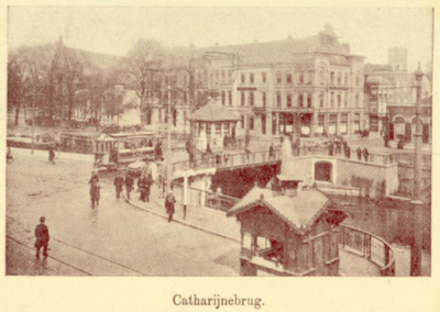 Ein altes Foto einer Stadtstraße mit Menschen, umgeben von Gebäuden, Bäumen, Pfählen und einer Brücke, mit Text unten.