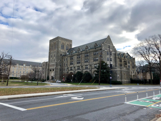 Großes Gebäude mit vielen Fenstern, als Universität von Pennsylvania etikettiert, umgeben von Bäumen, Straßenlaternen, Verkehrszeichen und Fahrzeugen an einem bewölkten Tag.