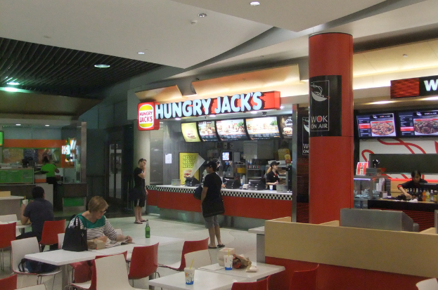 In einem Restaurant stehen Frauen an einer Essensbestellung, während an der anderen Seite Tische und Stühle mit einer Frau und einem Mann an einem der Tische angeordnet sind.