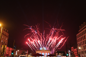 Eine belebte Straßen Szenerie in Berlin an Silvester, voller Menschen, Fahrzeuge und Gebäude, erleuchtet von Feuerwerk und Gebäudelichtern, die eine festliche Atmosphäre schaffen.