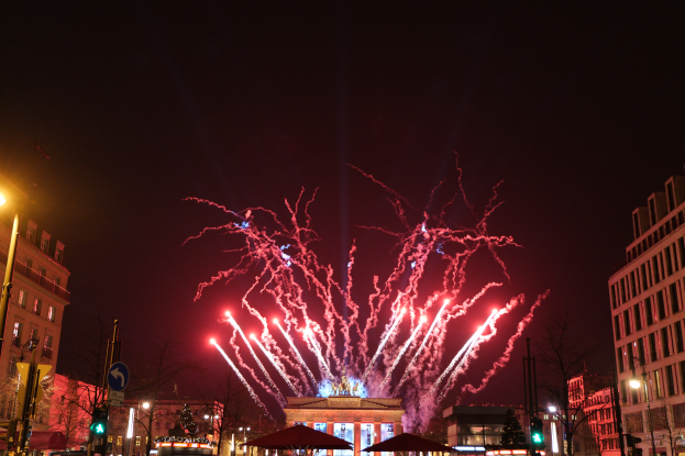 Eine belebte Straßen Szenerie in Berlin an Silvester, voller Menschen, Fahrzeuge und Gebäude, erleuchtet von Feuerwerk und Gebäudelichtern, die eine festliche Atmosphäre schaffen.