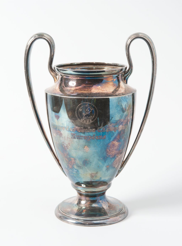 Der UEFA Champions League Pokal, ein stahlerner Becher mit zwei Händeln und einem blauen und silbernen Farbschema, mit der Inschrift «UEFA Champions League», vor einem weißen Hintergrund.