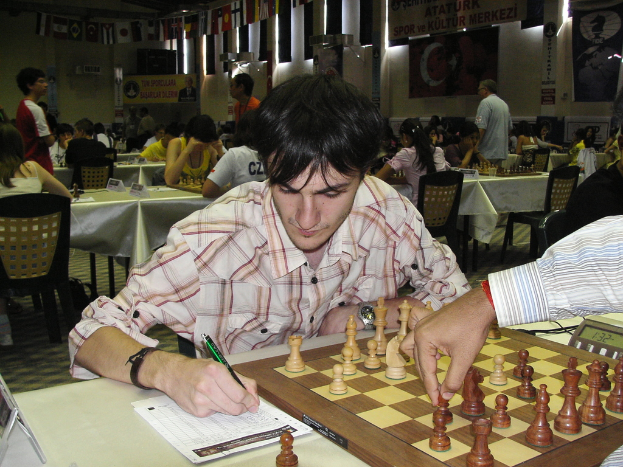 Eine Männerhand bewegt ein Schachstück auf einem Brett, während sie mit einem Stift schreibt, mit spielenden Menschen im Hintergrund.