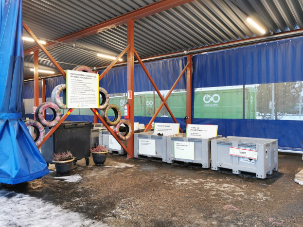 Großes Lagerinnere mit verschiedenen Reifen, Containern, Topfpflanzen, bedruckten Platten, Pfählen, Beleuchtung, einem blauen Tuch und anderen Gegenständen, umgeben von einer verschneiten Outdoor-Umgebung mit Bäumen und Fahrzeugen.