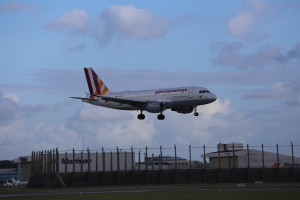 Germanwings Airbus A320-200 startet von Frankfurt Airport mit Gras darunter, einem Zaun im Vordergrund, Gebäuden und Bäumen im Hintergrund und einer bewölkten Himmel