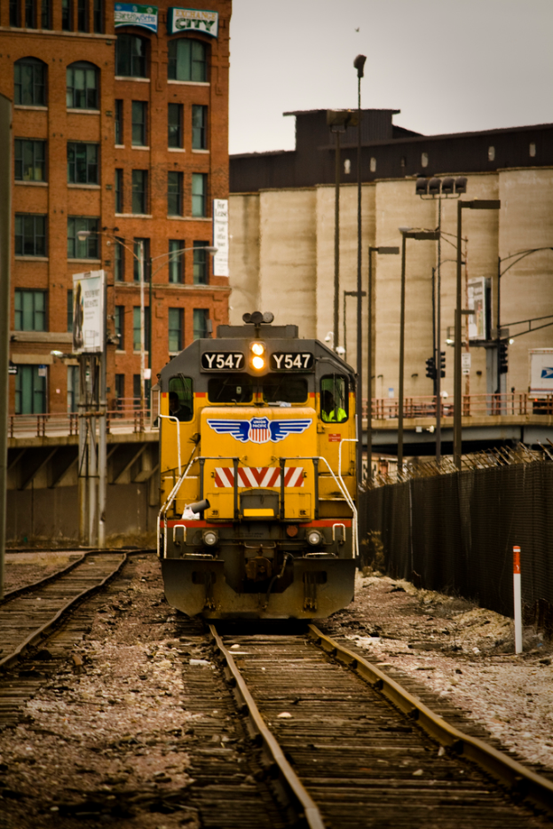 Ein Zug auf Schienen neben einem Zaun, mit Gebäuden und Himmel im Hintergrund.