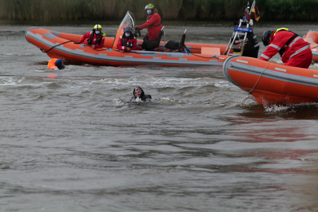 Eine Gruppe von Menschen in einem aufblasbaren Boot auf einem Fluss, mit zwei Personen im Wasser im Vordergrund und Vegetation im Hintergrund; alle tragen Schwimmwesten und Helme, was auf einen Rettungseinsatz hinweist.